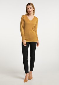 Mosterdkleurige v-halstrui van zachte brei, met lange mouwen en ribgebreide manchetten; gecombineerd met slanke zwarte broeken en beige puntige hakken.