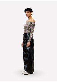 Vrouw met kort, krullend haar, gekleed in een off-shoulder patroon top, hoge zwarte leren broek met wijde pijpen, en zilveren loafers, staande opzij.