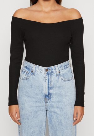 Femme portant un haut noir à manches longues épaules dénudées et un jean en denim bleu clair taille haute, debout sur un fond uni.