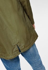 Person som bär en olivgrön jacka med en slits bak och blå jeans, visad från midjan till låren mot en neutral bakgrund.