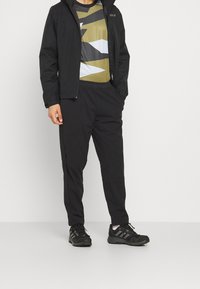 Mies, joka on pukeutunut mustaan takkiin, geometrisesti kuvioliseen paitaan, mustiin jogger-housuihin ja mustiin lenkkareihin, seisoo jalka toinen edessä ja toinen takana, toinen käsi taskussa.