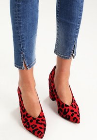 Zapatos de tacón alto con estampado de leopardo rojo, hechos de material suave, con punta afilada y tacón grueso, combinados con vaqueros azul cortos.