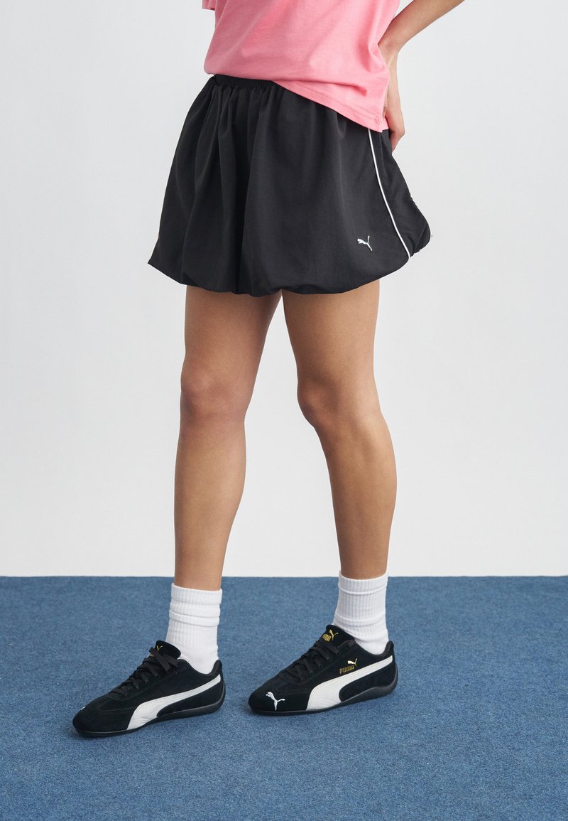 Personne portant une jupe de sport noire Puma, des chaussettes blanches et des baskets noires Puma, debout sur un tapis bleu avec les mains sur les hanches.