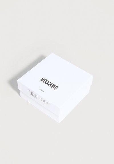 Λευκό τετράγωνο κουτί με μαύρο κείμενο "MOSCHINO Milano" στην κορυφή και μια μικρή ετικέτα προϊόντος στην πλευρά, τοποθετημένο σε ανοιχτή επιφάνεια.