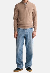 Beige Zip-Up-Pullover aus weichem Material, kombiniert mit hellblauen weit geschnittenen Jeans und schwarzen Sneakern mit Gummisohlen.