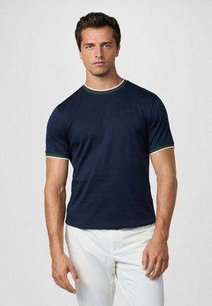 Hombre con camiseta de manga corta azul marino con ribetes verdes y blancos, combinado con pantalones blancos, de pie frente a un fondo claro.