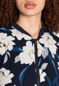 Blommig zip-up jacka med marinblå bakgrund, som har stora vita och pastellfärgade blommor, bladmönster och en ribbad krage.