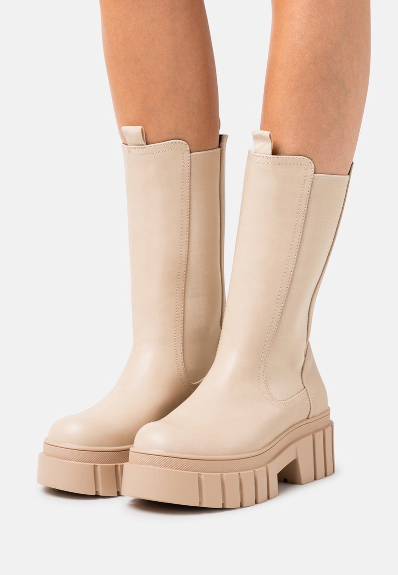 Bottes en cuir beige montant jusqu'aux genoux, dotées de semelles plateformes épaisses à rainures, bouts arrondis et languettes pour les enfiler, portées sur des jambes nues.