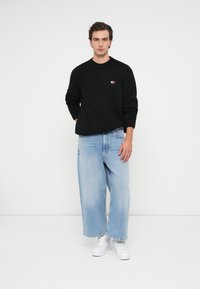 Czarny sweter dzianinowy z okrągłym dekoltem, w połączeniu z jasnoniebieskimi szerokimi spodniami dżinsowymi. Model nosi białe sneakersy; małe logo na piersi swetra.