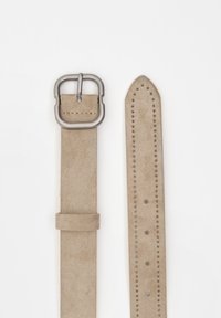 Beige velúr öv téglalap alakú fémbuckléval. Az övrészek több lyukkal rendelkeznek és sima felületük van, az öv pedig hegyes végződéssel keskenyedik.