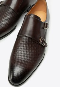 Dunkelbraune, strukturierte Leder-Mönchriemenschuhe mit doppelten silbernen Schnallen und spitzer Zehenform auf weißem Hintergrund.