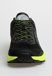 Black athletic shoe with neon green accents, textured upper, lace-up design, and rubber sole. Features branding on the tongue and side.