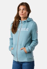 Lichtblauwe rits-hoodie gemaakt van een zachte katoenmix, met voorzakken, een trekkoordcapuchon en het woord "SEASIDE" in witte letters gedrukt.