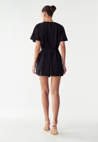 Črn romper s kratkimi rokavi in prilegajočim pasom ter gubano krilom. Narejen je iz gladkega materiala, ima neopazno zadrgo na hrbtu in je dopolnjen z nude petami.