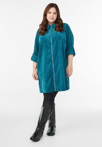 Sukienka w kolorze turkusowym z zamkiem z przodu, wykonana z miękkiego, teksturowanego materiału; posiada podwijane rękawy i luźny fason, połączona z czarnymi wysokimi botkami.