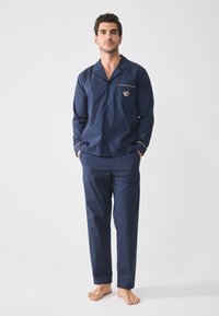Navy blue puuvillapaita yöasusetti, jossa on napilla suljettava paita, jossa on brodeerattu emblem ja kontrastivärinen reunus; suorat lahkeet, joustava vyötärö.
