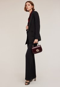 Zwarte op maat gemaakte blazer en uitlopende broeken met een kastanjebruine shirt. Model houdt een bordeaux handtas vast met gouden hardware; textuur hakken.