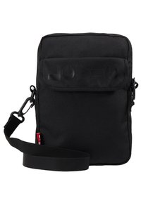 Černá látková crossbody taška s lesklým logem na přední straně, nastavitelným popruhem a zipovým uzavřením. Jednoduchý obdélníkový design.