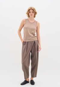 Bézs ujjatlan felső, texturált anyagból, lazán illeszkedő, taupe színű nadrággal, amelyen redők találhatók. A modellen fekete slip-on cipők vannak. Fehér háttér.