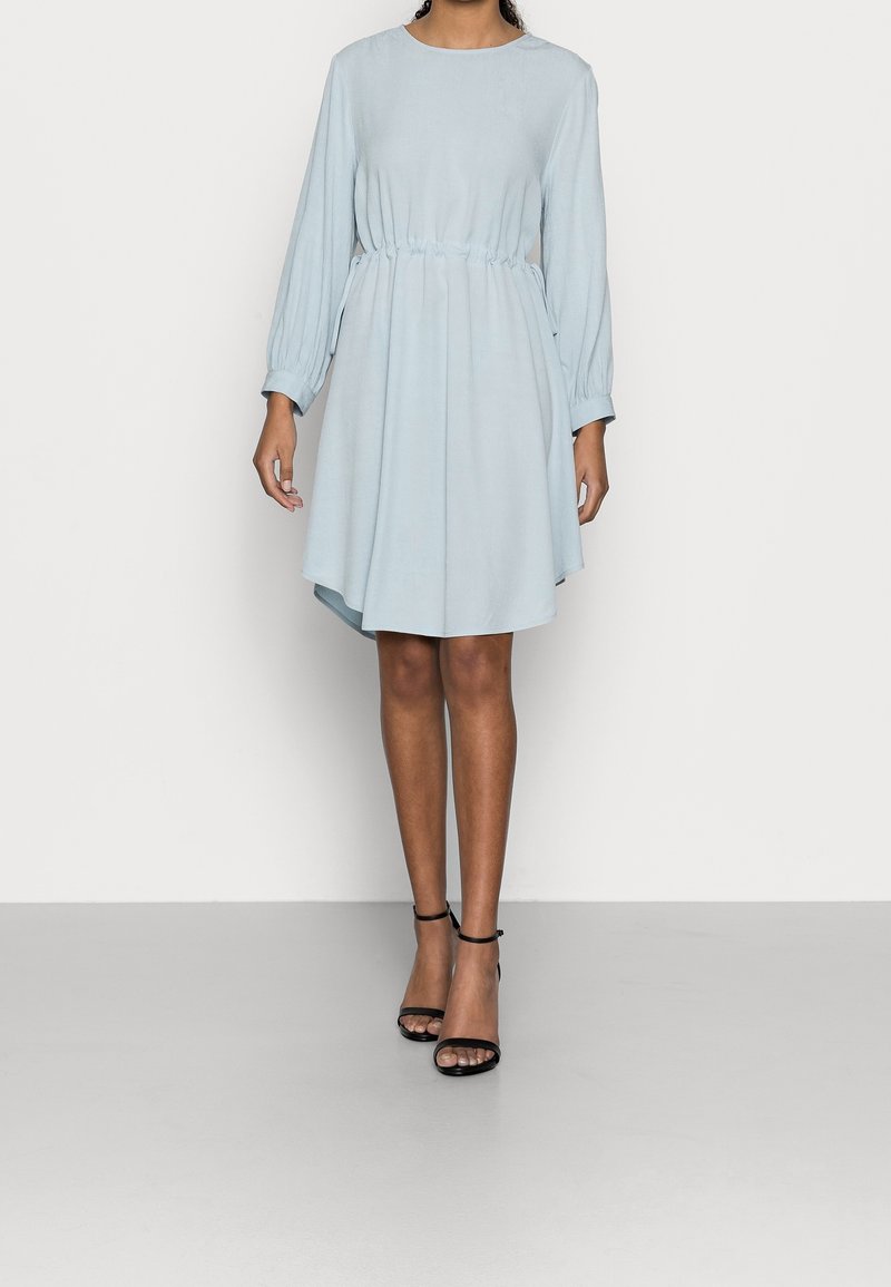 Femme portant une robe bleu clair à manches longues, longueur genou, avec taille froncée et sandales noires à talons hauts et bride à la cheville, debout sur un fond uni.