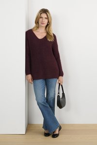 Burgunderfarbenen Strickpullover mit V-Ausschnitt und strukturiertem Muster, kombiniert mit ausgestellten blauen Jeans und schwarzen Ballerinas, während sie eine schwarze Handtasche hält.