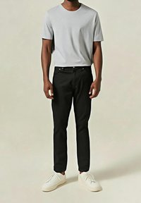 Homme debout portant un t-shirt gris clair à manches courtes, un pantalon noir et des baskets blanches sur un fond clair uni.