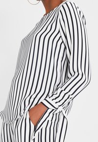 Blusa a righe bianca e blu navy, con maniche lunghe adornate da dettagli legati. Realizzata in tessuto morbido, presenta righe verticali su tutta la superficie.