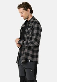 Zwart en grijs geblokt flanellen overhemd met knoopsluiting, opgerolde manchetten en een borstzak, gedragen over een zwart T-shirt.