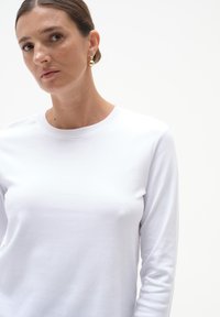 Biała bluzka z długim rękawem, wykonana z miękkiego materiału, z okrągłym dekoltem i dopasowanym krojem. Modelka uzupełniła stylizację dużymi złotymi kolczykami.