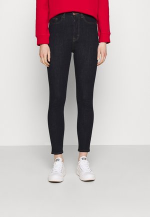 Frau trägt dunkle Skinny-Jeans, weiße Sneakers und ein rotes Langarmshirt, steht vor einem schlichten hellen Hintergrund.