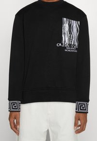Felpa nera con maniche lunghe, dotata di una tasca sul petto a fantasia, dettagli in testo bianco e accenti geometrici a contrasto sui polsini.