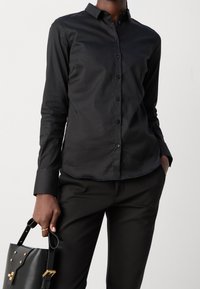 Camicia nera a maniche lunghe con bottoni, realizzata in tessuto morbido, con colletto classico e design aderente, abbinata a pantaloni neri opachi e una borsa.