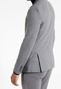 Szary wełniany blezer o dopasowanym kroju, z klapami z nacięciem i mankietami na trzy guziki. Z tyłu posiada pojedyncze rozcięcie i minimalną ilość detali.