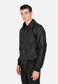 Giacca bomber nera in pelle con colletto, chiusura a bottoni, tasche laterali e polsini a costine. Texture liscia, design aderente.