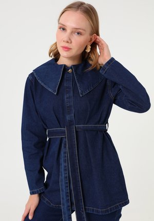 Lyshåret kvinde, der rører ved guldringe, iført mørkeblå denimjakke med bælte og bred, spids krave mod en ensfarvet baggrund.