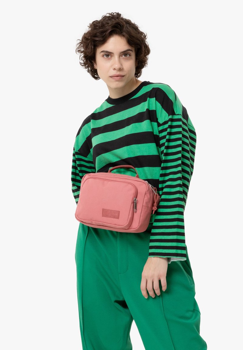 Rosa, rektangulär crossbody-väska med dragkedja och handtag på toppen, buren av en modell i en grön och svart randig skjorta och gröna byxor.