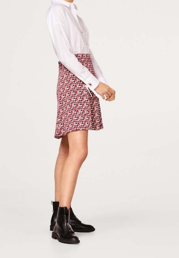 Femme portant une chemise blanche à manches longues, une jupe rouge à motifs et des bottines noires, debout de profil sur un fond clair uni.