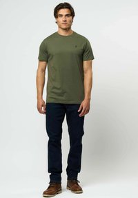 Jaunas vyras stovi, vilkėdamas alyvuogių žalia trumpomis rankovėmis marškinėlius, tamsiai mėlynas džinsus ir rudus odinius batus, ant paprasto fono.