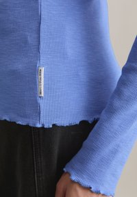 Geripptes Langarmshirt in Hellblau mit welligem Saum und Bündchen. Verfügt über ein kleines Etikett an der Seite. Das Material wirkt weich und dehnbar.