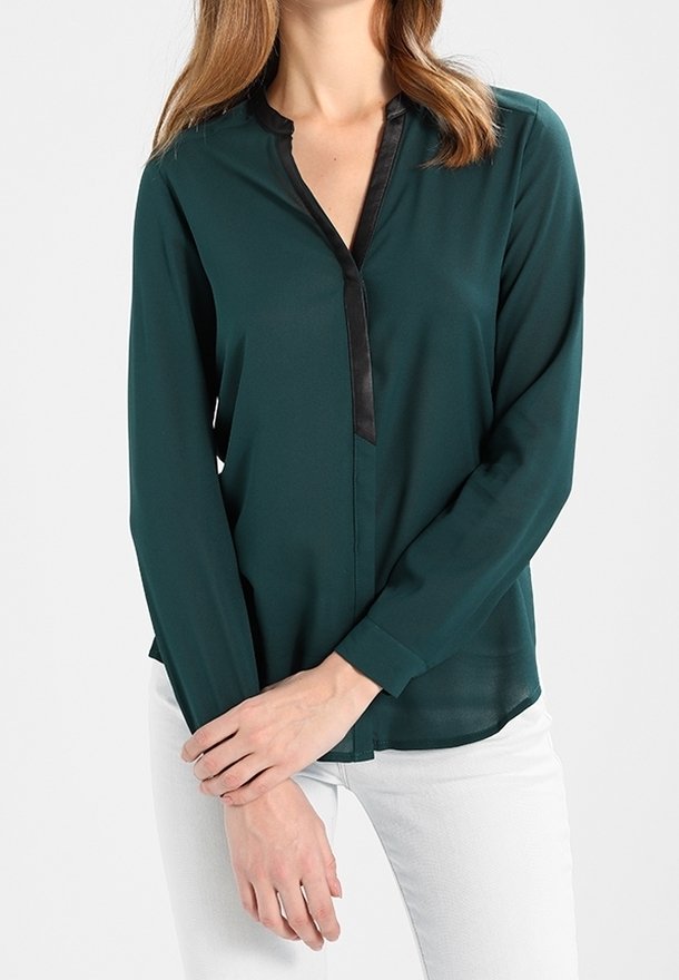 Femme portant un chemisier vert foncé à manches longues avec liseré noir et un pantalon blanc, debout les mains croisées devant elle.