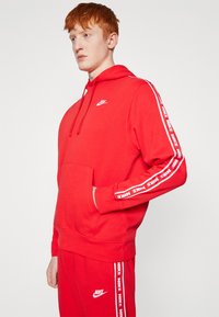 Rdeča Nike kapuca z kenguru žepom, nastavljivim kapuco na vrvico in belim logotipom. Ima bele stranske črte z napisom "NIKE".