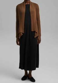 Ribbelachtige bruine cardigan met een open voorkant, gelaagd ontwerp dat over een zwart jurk tot de enkels valt, gecombineerd met zwarte instap schoenen.