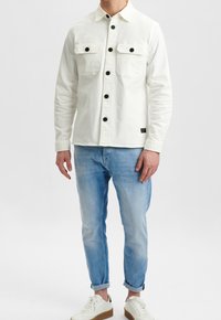 Giacca bianca fitted con due tasche sul petto, bottoni neri e maniche lunghe, abbinata a jeans blu chiaro arrotolati e sneakers bianche.