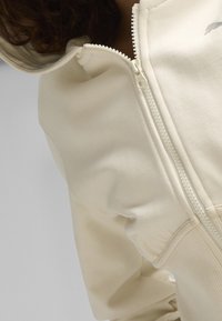 Beige zip-up hoodie met een gladde textuur, voorzien van een hoge kraag, een aansluitend ontwerp en een contrasterende rits. Minimalistische branding op de borst.