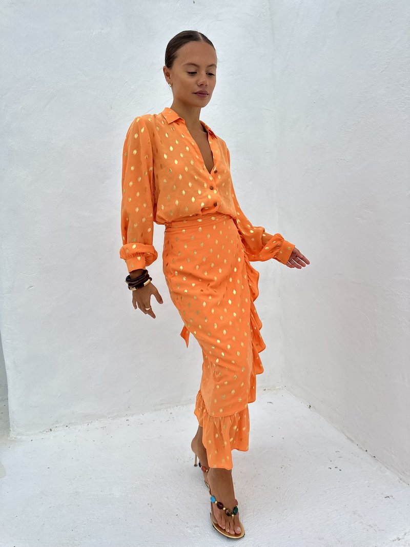 Robe en soie orange avec des pois dorés ; manches longues, col polo et ourlet à volants. Associée avec des sandales à lanières multicolores.