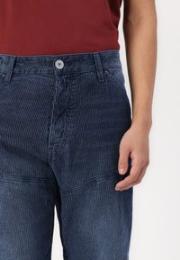 Blue vertically striped denim pants feature a button closure, side pockets, and a fitted waist. The fabric has a textured finish.