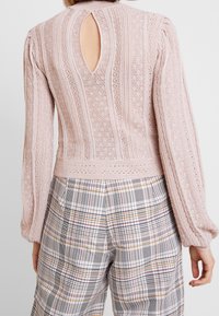 Jasnoróżowy sweter dzianinowy z pionowymi paskami, detalem w formie wycięcia z tyłu i teksturowymi wzorami, w połączeniu z spodniami w kratę.