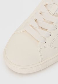 Sneaker bianco con tomaia sintetica liscia, punta rotonda, lacci piatti e suola in gomma testurizzata con dettagli di cucitura minimi.