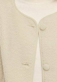 Gros plan d'une veste en laine beige texturée avec des boutons recouverts de tissu sur un haut en maille blanc.