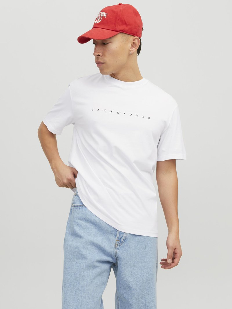 Weißes T-Shirt mit kurzen Ärmeln und dem Aufdruck "JACK & JONES" in Schwarz auf der Brust, kombiniert mit hellblauen Jeans und einer roten Baseballmütze.